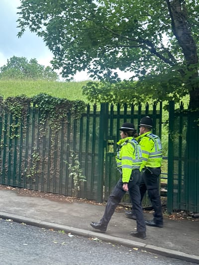 The image displays two uniformed British police officers, a male and a female, walking from left to right on a paved footpath in Epsom and Ewell, United Kingdom. The female officer, positioned to the left, wears a dark, brimmed police hat, a bright yellow high-visibility jacket with reflective silver stripes, dark trousers, and black boots. The male officer, positioned to the right and slightly behind, wears a traditional black British police custodian helmet, a matching bright yellow high-visibility jacket with "POLICE" visible on the back, dark trousers, and black boots, with a black equipment holder attached to his belt. Their action is a coordinated walk along the path. Immediately to their right, running parallel to the footpath, is a tall, dark green metal fence, partially obscured and heavily overgrown with green ivy and other foliage. Beyond the fence, a gentle green grassy slope ascends, topped by several trees under a bright, overcast sky. A section of a dark grey asphalt road is visible in the immediate foreground.