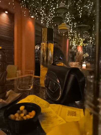 The image depicts an intimate, dimly lit indoor scene, likely a cafe, bar, or restaurant, taken from a low angle across a polished, dark table. The setting is characterized by warm, artificial light, primarily from numerous small, twinkling string lights suspended overhead, possibly intertwined with greenery, and diffuse orange lighting from walls and distant fixtures.

The main subjects on the table include a tall, clear bottle filled with a golden-yellow liquid, an empty clear glass tumbler, and a small black purse or bag with a prominent silver 'D' shaped logo, suggesting the brand Diesel. Scattered across the table are several yellow paper receipts, one visibly marked "epay" and another displaying a QR code. A small black bowl on the left contains various colorful, coated snacks or candies. Part of a dark, patterned object, possibly a decorative mug, is also seen on the far left.

In the soft-focus background, orange-colored structural elements and other hanging lamps are visible, contributing to the cozy atmosphere. The activity appears to be a casual evening gathering, with food and drink items suggesting a social occasion. The time of day is clearly night or late evening due to the reliance on artificial lighting. The location is Palaio Faliro, Greece, implying a local establishment. The overall mood is relaxed and inviting, with no people directly visible in the frame.