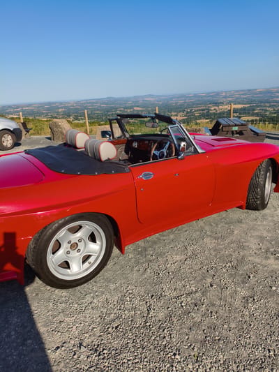 A bright red convertible sports car is parked on a gravel surface with a panoramic view of a rural landscape under a clear blue sky. The car is positioned at an angle, with its driver's side facing the camera. The interior features two headrests with grey and red accents. The car's black soft-top is partially deployed. In the background, a wooden picnic table and a fence are visible, separating the car from the rolling hills and fields of the countryside. The location is Cleehill, United Kingdom.