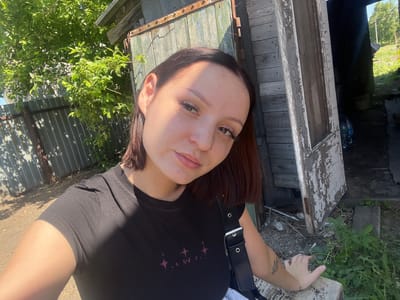 A young woman with dark hair and freckles takes a selfie outdoors in a rural area, possibly Zudilovo, Russia. She is wearing a black t-shirt and leaning against a weathered wooden structure with a green door. Sunlight casts shadows on her face and the surrounding environment. To the left, a metal fence and lush green foliage are visible, while to the right, the entrance to a dark interior space is partially obscured by the door and shadows. The ground is a mix of dirt and gravel, with some scattered debris. A black strap from a bag is visible across her chest, and a tattoo can be seen on her forearm. The overall scene suggests a casual, unposed moment in a rustic setting.