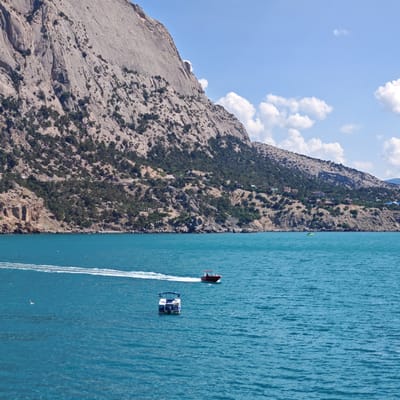 The image depicts a coastal scene with a large, rocky mountain dominating the background. The mountain is covered with sparse vegetation and shows signs of erosion. In the foreground, the turquoise blue sea is calm, with ripples indicating a gentle breeze. Two boats are visible on the water. One is a small red motorboat moving at speed, leaving a white wake. The second boat is a white pontoon boat with a blue canopy, stationary in the water. The shoreline is lined with trees and the base of the mountain. The sky is a clear blue with scattered white clouds. The location is Novyi Svet, Ukraine, a town known for its picturesque coastline and mountainous terrain.