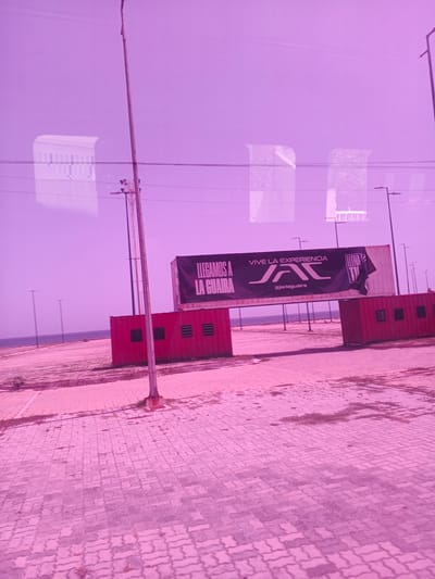 This media file captures an outdoor scene in Macuto, Venezuela, under what appears to be a bright, possibly overcast sky. The dominant color cast is a vibrant pink, suggesting an unusual lighting condition or photo editing.
The foreground is paved with interlocking bricks, showing some patches of dry grass. The midground features two large, red shipping containers placed side-by-side, forming an entryway. A black banner is stretched across the top of these containers. The banner displays text and a logo. The text reads "LLEGAMOS A" on the left, and "VIVE LA EXPERIENCIA" on the right, above a stylized logo that appears to be "JAC". Below the logo, the text "@jaclaguaira" is visible. A portion of the banner on the far right is ripped and folded.
Beyond the containers, a vast, flat, sandy area stretches towards the ocean, which is visible as a thin blue line in the distance. Several tall, thin poles, likely streetlights or flagpoles, are scattered across this area and further into the distance. Reflections of buildings, possibly a grand structure with columns and arched windows, are visible in the upper portion of the image, suggesting the photograph was taken from inside a vehicle with a window.
The scene appears to be set up for an event or as an entrance to a venue. The overall mood is somewhat surreal due to the strong pink hue, but the setting itself is one of an open, coastal area with commercial or event infrastructure. There are no people visible in the frame. The time of day is unclear due to the uniform lighting and pink cast, but the bright sky suggests daylight.