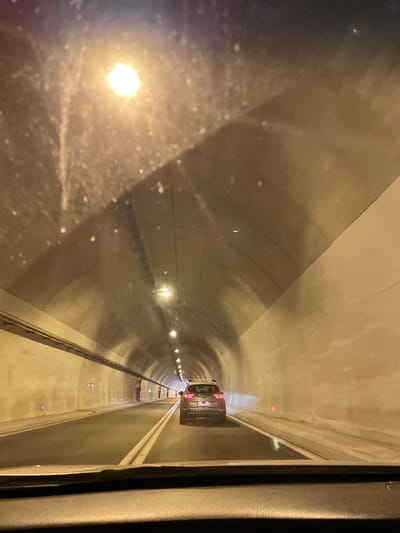 The media file shows the interior of a tunnel, likely a road tunnel, as evidenced by the presence of cars and lane markings. The tunnel is illuminated by artificial lights, suggesting it is nighttime or that the tunnel lacks natural light. The overall color palette is dominated by grays and yellows, consistent with concrete structures and artificial lighting.
In the foreground, a significant portion of the image is taken up by what appears to be a reflective surface, possibly a window or a windshield. This surface is dirty or smudged, with visible specks and streaks, and it reflects a bright light source, possibly an external light or a light within the tunnel.
Further into the tunnel, several vehicles are visible, traveling in the same direction. One car, in particular, is clearly seen, with its rear lights illuminated. The car appears to be a passenger vehicle, and its license plate is partially visible, showing the characters "CG" followed by other characters that are not fully legible. Red marker lights are also visible along the tunnel ceiling.
The tunnel walls are made of concrete and have a curved, arched shape, characteristic of tunnel construction. There are also railings or barriers visible along the sides of the tunnel. The scene conveys a sense of movement and transit through an enclosed space. The location is identified as Komotini, Greece. There are no discernible people or emotional cues in the image. The weather is not apparent.