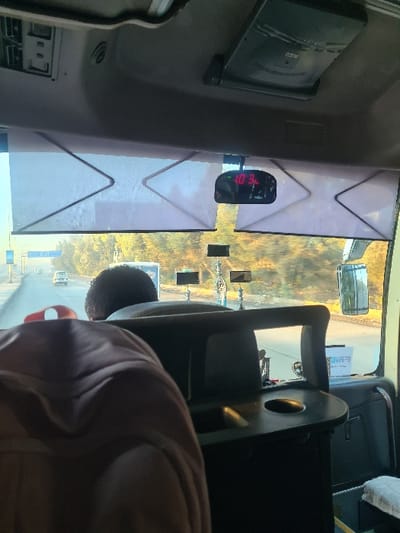 Inside a vehicle, possibly a bus, the scene is oriented in landscape view.  A black seat is in the foreground. A passenger is visible in the bottom right. A digital clock reads "10:34". White and black window shades are present, partially obscuring the outside view of trees. A mirror is seen in the upper-right corner of the window, reflecting the outside scenery. A small television screen is visible in the upper-left corner.