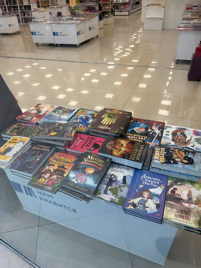 The image shows a display of books in a store in Novoalexandrovka, Russia. The books are arranged on a white shelf or box. The covers of several books are visible, including "ДНЕВНИК 3" (Diary 3), "ХОДЯЧИЙ ЗАМОК" (Howl's Moving Castle), "ХРОНИКИ НАРНИИ" (The Chronicles of Narnia), "КОРАЛИНА" (Coraline), "Девочка с Лисьим Хвостом" (The Girl with a Fox Tail), "ДЕВОЧКА-КАПИТАН" (Girl-Captain), "Приключения кота-детектива" (Adventures of the Cat-Detective), "ОХОТНИКИ ЗА МИФАМИ" (Myth Hunters), "Мейзи Хитчинс", "Призрак Луна-Парка" (The Ghost of the Moon Park). The phrases "НАМ НРАВИТСЯ" (We Like It) are printed on the sides of the display. The floor is tiled. Other shelves of books are visible in the background.