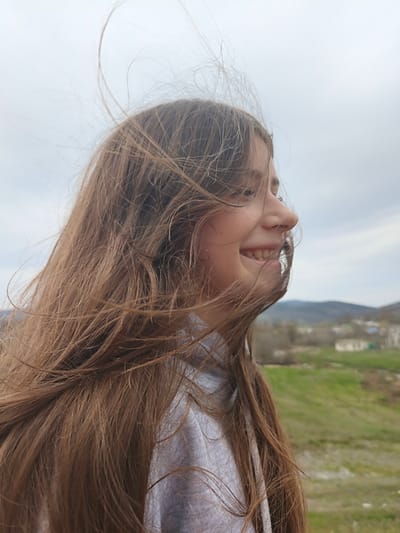A young woman with long, light brown hair stands outdoors, her hair being blown by the wind. She is smiling and looking towards the right side of the frame. The background includes a green, grassy field, some buildings, and hills or mountains in the distance. The sky is overcast. The location appears to be in a rural or semi-rural area. The exact location within Орлиновский округ, Ukraine cannot be determined from the image.