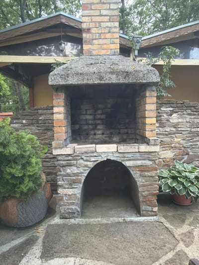 The image captures an outdoor barbecue and fireplace structure crafted from a combination of brick, stone, and concrete. The central feature is a rectangular firebox constructed from multi-toned bricks, showing signs of use with darkened interior walls. A thick, grey concrete slab forms the roof over the firebox, extending upwards as a chimney base. To the right, an arched opening is integrated into a rough-hewn stone and brick wall, providing a niche or secondary cooking area.

Green foliage surrounds the structure. A potted Hosta plant with variegated leaves is placed on a stone shelf to the right of the arched opening. Below, a dense green conifer or shrub thrives near the base, alongside a textured, spherical garden ornament in grey and brown. In the background, elements of a building are visible, including wooden eaves, a dark reflective surface (possibly a window or panel) with a reflected outdoor light fixture, and a portion of a roof. Mature trees with lush green leaves are seen beyond the building.

The ground surface consists of irregularly shaped grey flagstones, forming a patio area. The lighting suggests it is daytime, with soft, diffused light. There are no individuals present, and no specific activities are taking place. The scene depicts a serene and well-established outdoor space, set in Sushina, Bulgaria. No visible text is present in the image.