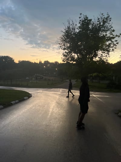 The image, taken in Bukovlak, Bulgaria, depicts an outdoor scene at dawn or dusk, characterized by a gradient sky transitioning from orange-yellow at the horizon to pale blue-grey overhead, with indistinct cloud formations. A wet, paved road occupies the foreground and mid-ground, exhibiting reflective streaks from ambient light. To the far left, a patch of green grass is bordered by a curb featuring black and white markings. Two silhouetted individuals are visible on the road. In the foreground right, a person is partially visible from the waist down, wearing dark shorts and sneakers, standing on a skateboard. In the mid-ground left, another person walks from left to right, clad in dark clothing. Dense trees and foliage frame the scene, with a prominent leafy tree on the mid-right side. In the distant background, obscured by vegetation, are indistinct structures, a partially visible white vehicle, and illuminated streetlights or utility poles. The wet road surface indicates recent precipitation.