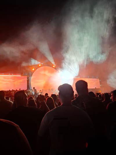 The media file captures a vibrant concert or festival scene, with a large crowd of people silhouetted against a brightly lit stage. The stage features a large circular screen displaying an orange, cloud-like pattern, resembling a sunset or fiery sky. Several spotlights emit beams of white and green light through a haze of smoke, creating dramatic atmospheric effects. The crowd, packed together and looking towards the stage, appears engaged and excited, though their faces are obscured by shadow.

The lighting suggests the event is taking place at night or in a darkened venue. The overall mood is energetic and immersive, characteristic of a live performance. Some individuals in the crowd are holding up their phones, likely to record or photograph the event. The image is oriented vertically, with the crowd occupying the lower portion and the stage dominating the upper half.

A small red and yellow logo, possibly a sponsor, is visible on the upper right side of the stage structure. The scene conveys the electrifying atmosphere of a public gathering centered around music or entertainment.

Location Context: Mala tsarkva, Bulgaria.