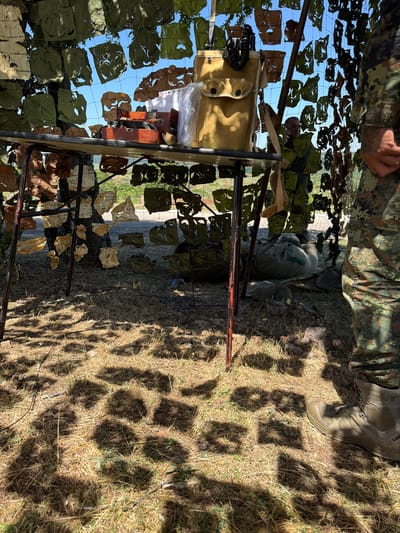 The image depicts an outdoor scene, likely a military training exercise, with a camouflage net draped over a structure. Beneath the netting, a table is set up with various objects, including what appears to be a radio or communication device, papers, and a canvas bag. A person in military camouflage uniform stands to the right, their legs and boots visible. In the background, another individual in camouflage is partially visible, engaged in a conversation. The ground is covered in dry grass, and shadows of leaves are cast upon it, suggesting a sunny day. The overall environment appears to be a rural or semi-rural area, possibly a field or training ground. The exact location in the city of Momin Sbor, Bulgaria, cannot be confirmed from the image alone, but the presence of military personnel and equipment suggests a training or operational context.