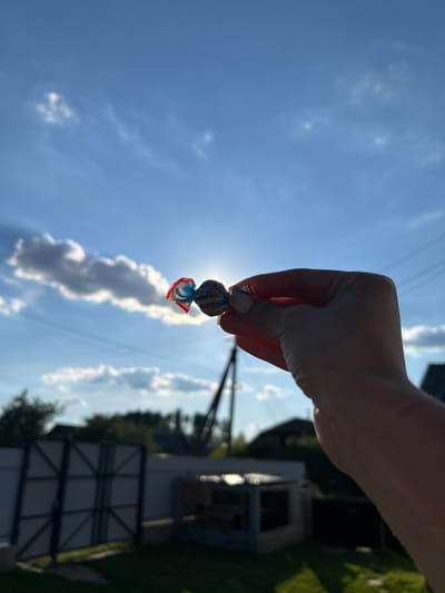 A hand holds a wrapped candy in front of a bright sky. The candy wrapper is red, white, and blue, and the candy appears to be spherical. The background is out of focus but shows trees, a fence, and some buildings. The image was taken outdoors during the day, with the sun positioned behind the candy, creating a lens flare effect. The general setting appears to be a rural or suburban area, possibly in Vyshevychi, Ukraine, given the style of the fence and buildings. The sky is blue with scattered white clouds.