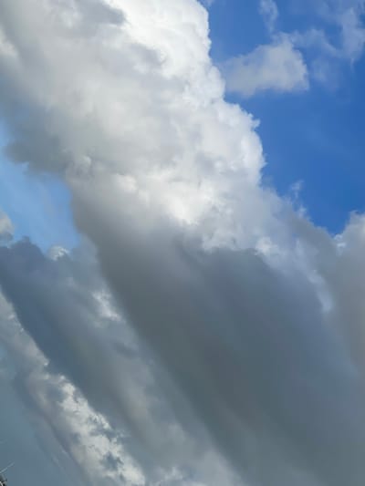 The image displays an outdoor scene, specifically the sky above the city of De Lier, Netherlands. The frame is predominantly filled with cloud formations against a backdrop of blue sky. Bright white, voluminous cumulus-type clouds are present in the upper-left and central-right portions. Extending diagonally across the image from the lower-left to the upper-right are darker gray, elongated cloud masses, exhibiting varying densities and a stratified texture. Patches of clear blue sky are visible in the upper-right quadrant and as an irregular shape in the mid-left. No individuals, discernible man-made structures, or objects are present within the visible frame. No actions or interactions are depicted.