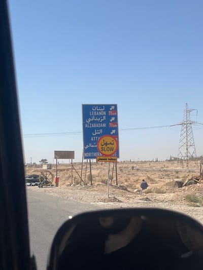 An outdoor daylight scene, likely viewed from a vehicle due to a partial dark frame on the left and a rearview mirror in the foreground, depicts a semi-arid environment in the City of Adra labor, Syria. The sky is clear and bright blue.
In the mid-ground, a prominent blue road sign with white text indicates directions and distances: "LEBANON 71Km" (right arrow), "ALZABADANI 71Km" (right arrow), and "ATTAL" (right arrow), along with corresponding Arabic script. Below it, a yellow circular sign reads "SLOW" (Arabic: تمهل), with additional Arabic text "شمالي" (Northern) and "نقطة تفتيش" (checkpoint). To the left of this sign, a smaller, blank rectangular wooden sign is mounted on posts. Further left, a large wooden spoked wheel rests near coiled black pipes or cables, and a red object, possibly a fire extinguisher, is also present.
The ground consists of reddish-brown earth with sparse vegetation and scattered rocks. A paved road surface is visible in the lower left. In the right mid-ground, a lone individual, dressed in dark clothing, walks away from the camera across the open, unpaved terrain. In the far right background, a large metal electricity pylon stands against the horizon. Debris and construction materials are scattered on the right side of the scene.