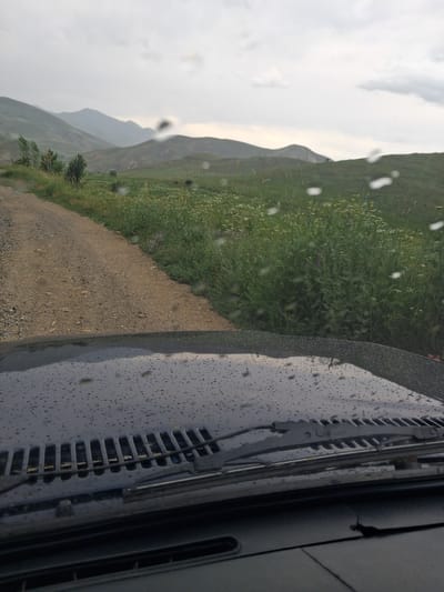 The image shows a view from inside a vehicle looking out at a rural landscape in Martiros, Armenia. The foreground is dominated by the dark, wet hood of the vehicle, covered in raindrops, and the windshield wipers are visible. Beyond the vehicle, a dirt road curves to the left, flanked by green vegetation and scattered wildflowers. Rolling hills and mountains, shrouded in a hazy, overcast sky, form the background. The weather appears to be cloudy and possibly rainy, as indicated by the water droplets on the windshield and the subdued lighting. No people are visible in the image.