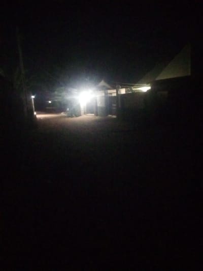 The image depicts a dimly lit outdoor scene, likely at night, with artificial lighting illuminating a building and its surroundings. The lighting sources appear to be bright, possibly from streetlights or exterior building lights, casting long shadows and creating a high-contrast environment. The building itself seems to be a residential or commercial structure, with visible windows and a roofline. Vegetation, such as trees or bushes, can be partially seen in the background and along the sides, suggesting an urban or suburban setting. The ground appears to be unpaved, possibly a dirt or gravel pathway. The overall impression is one of a quiet, dimly lit street or yard in Yola, Nigeria, with limited visibility due to the nighttime conditions and the quality of the lighting.
