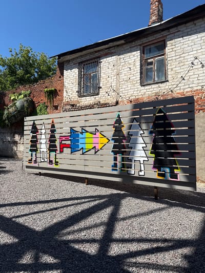 The image depicts a sun-drenched outdoor scene in the city of Yelets, Russia. In the foreground, a gray wooden fence is adorned with a colorful mural. The mural features stylized, silhouetted trees on the left, with outlines of bright neon colors at their bases. To the right of the trees, a large arrow composed of segments in various bright colors, including blue, green, yellow, pink, and red, points towards the right. The fence is set against a backdrop of a weathered brick building with white-washed bricks. Two windows are visible on the building's facade, one with metal bars and the other with a wooden frame. A string of lights is attached to the building's exterior. To the left of the building, a brick wall with some greenery and an unusual, large, moss-covered object is visible. The ground is covered in small gray gravel, casting distinct, elongated shadows from the fence and possibly unseen objects. The sky above is a clear, bright blue, indicating a sunny day.