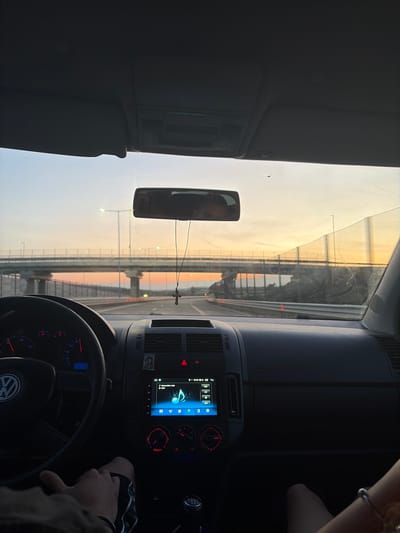The image depicts the interior of a vehicle, specifically a Volkswagen, from the driver's perspective, looking forward through the windshield at sunset. The location is Aldomirovtsi, Bulgaria.
The vehicle's interior features a steering wheel with a visible Volkswagen emblem on the left. The dashboard gauges, backlit in red and blue, indicate zero speed and RPM. Below the central air vents, an aftermarket infotainment system displays a music player interface showing a musical note icon, with the time "21:21" visible. Red-backlit climate controls are located beneath the display. A small religious icon is affixed to the dashboard above the infotainment unit. A manual transmission gear stick is visible in the lower center.
On the left side of the frame, the driver's hands and legs, wearing dark shorts, are partially visible. On the lower right, a portion of a passenger's leg or arm, adorned with a silver-colored bracelet, is visible. The rearview mirror, centrally located, has a crucifix pendant hanging from it and shows a distorted reflection of the driver's upper face. The car's sun visors and headliner are visible at the top of the frame. The windshield exhibits multiple scratches and smudges, particularly on the right side, and a small insect is present near the top center of the glass.
The exterior scene, viewed through the windshield, shows a highway at dusk or sunset. The sky transitions from light blue at the top to orange, pink, and yellow hues near the horizon. A large concrete overpass or bridge structure spans the highway ahead, with multiple support pillars. Streetlights are visible on and around the bridge, some emitting light. Roadside barriers and transparent or mesh sound barriers line the highway. Another bridge or elevated road structure is faintly visible in the far distance beyond the immediate overpass.