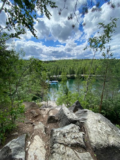 This is a high-angle shot of a rocky, wooded area overlooking a body of water. In the foreground, large grey rocks are scattered across the ground, with some vegetation growing between them. The path appears to be unpaved and leads down towards the water.
In the middle ground, the water is a deep green color, and several small boats are docked along the shore. The boats are light blue and white. Surrounding the water is a dense forest of green trees that extends to the horizon.
The sky above is a mixture of blue sky and white, fluffy clouds. The overall impression is one of a natural, serene landscape.
This scene is likely located in the Ruskeala Mountain Park in Ruskeala, Russia, a popular tourist destination known for its marble quarries and scenic beauty. The park features a large, flooded marble quarry that has been transformed into a picturesque lake.
