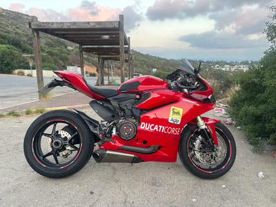 The image shows a red Ducati motorcycle parked on a paved surface next to a roadside. The motorcycle is a high-performance sportbike, featuring a sleek design with prominent branding, including "DUCATI Corse" and "Agip" stickers. The motorcycle has carbon fiber accents and an Akrapovič exhaust system.
In the background, there is a wooden structure that appears to be a covered seating area or shelter, possibly part of a rest stop. Behind the shelter, there is a sloping hillside covered with green vegetation. Further in the distance, a town or settlement with buildings is visible on the hillside, and beyond that, a hazy horizon suggests a view of the sea or a large body of water. The sky is partly cloudy with soft, diffused light, indicating it might be late afternoon or early evening.
The location is specified as the city of Mate Asher Regional Council, Israel. The overall scene depicts a motorcycle parked in a scenic, somewhat rural setting, likely along a road with a view. There are no people visible in the image. The ground around the motorcycle shows some litter, suggesting the area might be a public space.