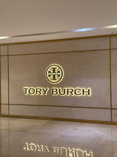 The image shows a vertical sign for "TORY BURCH" illuminated by warm lights. The sign is mounted on a wall that appears to be made of marble or a similar stone, with gold-colored trim. To the left of the text is the iconic Tory Burch logo, a circular emblem with a stylized double "T" inside. The lighting creates a soft glow around the lettering and logo, suggesting an indoor setting, possibly a retail store or a mall. The background wall has a subtle texture and pattern. On the far right of the image, reflected in the wall or a separate panel, are the letters "HOUB YROT", which appear to be a reversed or mirrored version of the "TORY BURCH" text. This suggests a polished surface or a display with mirrored elements. The scene is static, with no discernible activity taking place. The overall impression is one of modern and elegant branding. The text "TORY BURCH" is clearly legible. The location is identified as Xuanwu District, China.