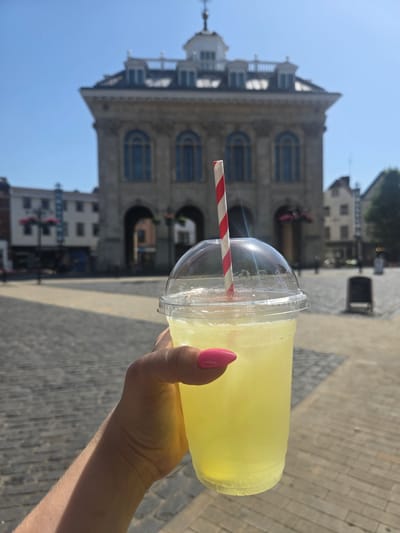 The image shows a person holding a clear plastic cup filled with a yellow beverage, likely lemonade. A red and white striped straw is inserted into the cup. The person's hand, with bright pink nail polish, is visible gripping the cup. The background suggests an outdoor setting with a cobblestone pavement and a large, ornate building with arched doorways and windows, possibly a town hall or historical landmark. The sky is clear and blue, indicating a sunny day. The building's architecture, along with the cobblestone, points towards a location in the Vale of White Horse, United Kingdom. The lighting and shadow play suggest it is daytime, likely mid-morning or afternoon. The overall impression is one of enjoying a refreshing drink on a pleasant day in a historical European town square. No visible text is present in the image.