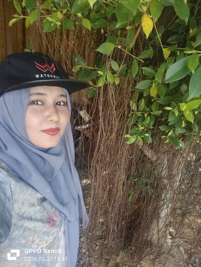 A woman wearing a blue hijab and a black baseball cap stands facing the camera. The cap has the word "WATCHOUT" on it. The woman is wearing a floral patterned garment. She appears to be standing in front of a wall of vines and leaves. The lower left corner of the image includes the text "OPPO Reno8 2026.05.23 15:51". The location is implied to be Santan, Indonesia.
