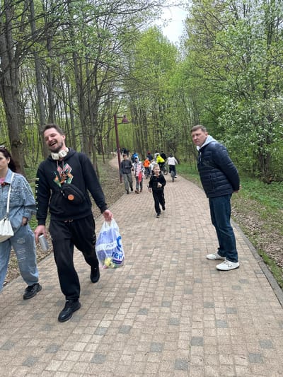 The scene shows a paved path in Grebnevo, Russia, lined with trees. A group of people are walking along the path. In the foreground, a man is holding a plastic bag and a can. Further down the path, people are walking and cycling. Another man is standing with his arms crossed. The trees are in bloom, suggesting it is springtime.