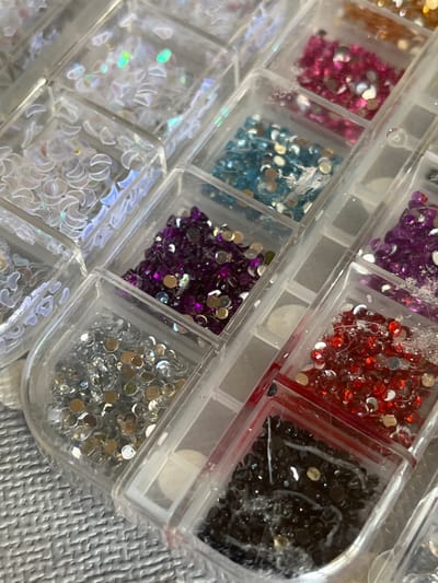 The image displays a close-up, overhead view of a clear plastic organizer filled with various colored rhinestones and nail art embellishments. The organizer is divided into multiple small compartments, each containing a different type of embellishment. In the upper left, there are white crescent-shaped embellishments with iridescent effects. Moving to the right, there are compartments filled with pink, blue, purple, red, and black rhinestones. Some compartments also contain smaller, round, metallic-colored beads. The surface beneath the organizer appears to be a textured, light-colored material, possibly a mat or table. The lighting suggests an indoor setting. There is no indication of the city of Figueres, Spain, or any specific location within a city. The image solely focuses on the collection of craft supplies.