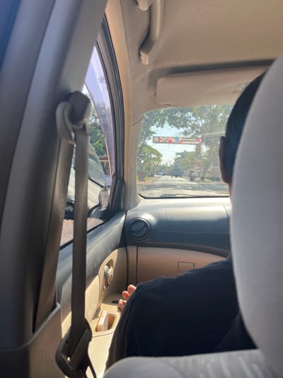 The image, taken from the interior of a vehicle, provides a view through the windshield and side window. The perspective is from the passenger seat, looking forward and slightly to the right. In the foreground, the interior of the car is visible, including the dashboard, a seatbelt, and a portion of the passenger's head and shoulder. The passenger is wearing a dark t-shirt. Outside the car, a street scene unfolds. Trees line the road, and buildings are visible in the distance. There is traffic on the road, including other vehicles and motorcycles. A large archway with signage spans the road in the middle distance. The environment suggests an urban or semi-urban setting. Based on the signage, the location is identified as Kedungreja, Indonesia. The lighting suggests it is daytime, likely sunny.