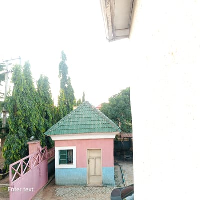 This is a photograph of a small pink and light blue building with a green tiled roof, situated in Azu, Nigeria. The building has a single tan-colored door and a green-paned window. In front of the building is a pink metal gate with an ornate lattice design. Tall, slender trees with dense green foliage line the background. The ground is paved with interlocking stones. A section of a white wall occupies the right side of the frame, and a sliver of a dark-colored car is visible in the lower right corner. The sky is bright and washed out, suggesting it is daytime. The overall scene depicts a residential or possibly a security post environment.