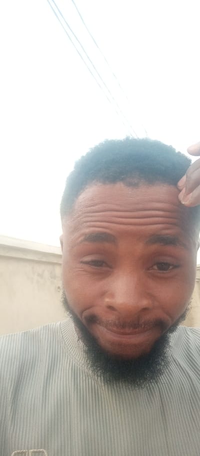 A close-up portrait of an adult male is captured in Tunga Mekidi, Nigeria. The subject, of apparent African descent, has short, dark, textured hair and a dark, neatly trimmed beard. He wears a light-colored, possibly pale blue or grey, ribbed short-sleeved garment with a mock turtleneck collar. His face shows a slight smile, with crinkling around his eyes. His left hand, visible on the upper right side of the frame, is positioned with fingertips near his right temple, appearing to touch his hairline. The background features a light-colored, possibly off-white or beige, textured wall on the left side, and a bright, largely featureless sky occupying the upper right, traversed by three thin, light-colored diagonal lines, consistent with power lines or wires.