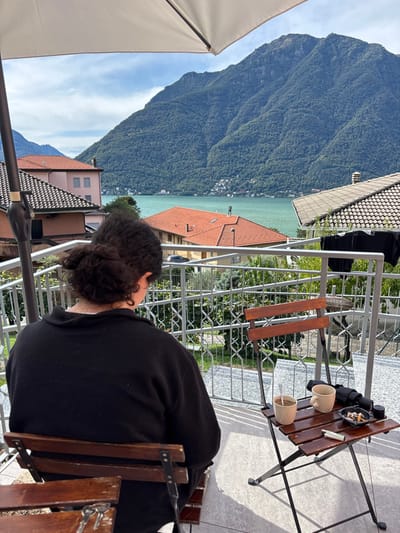 A single individual is seated with their back to the camera on an outdoor terrace in Nesso, Italy, overlooking Lake Como and forested mountains under a partly cloudy sky. The person has dark, curly hair tied back in a bun and wears a black long-sleeved top, sitting on a wooden slatted chair with metal supports. A set of keys rests on an adjacent wooden chair to the left in the foreground. To the right of the seated individual, a small wooden folding table with metal legs holds two light-colored ceramic mugs (one containing a spoon), a black ashtray filled with cigarette butts, a white cigarette, a black cylindrical object, and a folded dark fabric item. The terrace floor is gray tile, and a metal railing with geometric patterns separates the immediate area from the view. Beyond the railing, residential buildings with orange and gray tiled roofs are visible, descending towards the turquoise water of Lake Como. Across the lake, a distant settlement is discernible at the base of expansive, green, tree-covered mountains that dominate the background. A large white patio umbrella is partially visible in the upper left of the frame.