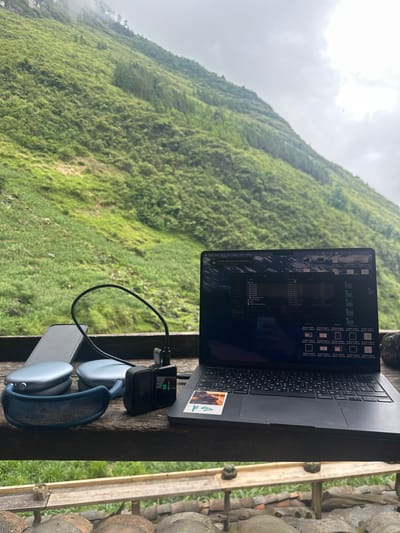 A laptop computer, possibly a MacBook, is open on a wooden surface with a view of a verdant, steep hillside in the background. The vegetation on the hillside appears dense and lush, possibly mossy, and partially obscured by mist or low clouds. To the right of the laptop is a pair of blue AirPods Max headphones resting on top of a grey smartphone and a black charging adapter with a short cable connected. The laptop screen displays a file explorer window with multiple files and folders. The wooden surface appears to be part of a structure with additional wooden elements to the right and stone materials in the background. The scene is located in the Yên Minh Commune, Vietnam.