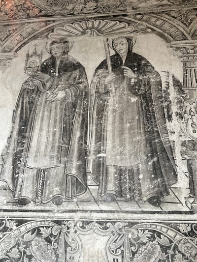 The image shows a mural painted on a wall, likely in Huejotzingo, Mexico. The mural depicts two robed figures standing side-by-side. The figure on the left is holding a monstrance in their left hand and a book in their right hand. They are wearing a hooded robe and have a halo around their head. The figure on the right is holding a staff or cross in their hands. They are also wearing a hooded robe and have a halo around their head. Both figures appear to be standing on a tiled floor, and there is decorative stonework above them. The mural is in black and white, and shows signs of wear and tear. The overall style suggests religious iconography, possibly depicting saints or religious figures. The lower part of the image shows a decorative border with floral motifs.