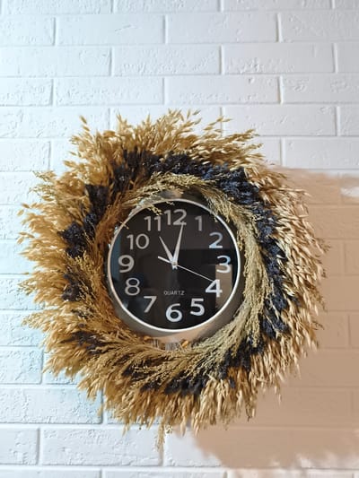 A round, black-faced clock with white numbers and hands is centered on a white brick wall. The clock is surrounded by a decorative wreath made of dried wheat stalks and dark purple sprigs, possibly lavender. The wreath is artfully arranged to frame the clock face. The lighting suggests an indoor setting, with the white brick wall showing texture and slight shadows. The scene is static, with no people or apparent actions. The location is identified as Zadebri, Ukraine.