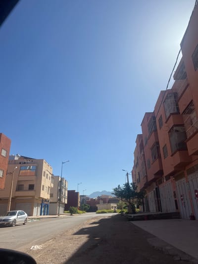 The image depicts a residential street scene in Beni Mellal, Morocco, under a clear, bright blue sky with the sun positioned high on the right, casting long shadows.
On the right side of the street, a continuous row of multi-story buildings, predominantly reddish-orange in color, extends into the distance. These structures feature multiple windows, many with metal grates or decorative cages, and some small balconies. The ground floors are characterized by large, closed metal roller shutters or garage-style doors, suggesting commercial or storage use. A utility pole with overhead wires is visible near the foreground.
On the left side, further back, are several multi-story buildings of lighter, beige or light brown coloration. A silver sedan, appearing to be a Seat Leon, is parked parallel to the curb in front of one of these buildings, which has blue-accented ground-floor shop fronts.
The street itself is paved asphalt, flanked by unpaved dirt shoulders on both sides, with the right-hand shoulder appearing wider and more uneven. Multiple modern streetlights are positioned along the street. A small tree with green foliage is present on the right shoulder. No individuals are visible in the scene. In the far distance, a range of barren mountains is silhouetted against the horizon. The bottom left corner shows a small portion of what appears to be a vehicle's interior.