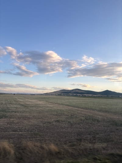 This is a vertical, eye-level, outdoor, landscape photograph taken in the city of Naiden Gerovo, Bulgaria. The scene depicts a rural area under a vast, blue sky filled with scattered, fluffy white and grey clouds. The lower half of the image is dominated by a dry, golden-brown field, suggesting it might be late summer or early autumn. The field appears to have been recently harvested or is fallow, with visible rows and patches of stubble. A dirt path or track runs diagonally across the field, leading towards the background. In the distance, rolling hills covered in dark green vegetation rise from the plain. Several structures, possibly houses or farm buildings, are scattered along the base of the hills, and two prominent cell towers are visible on the hilltops. The overall impression is one of tranquility and open space. There are no people or animals visible in the frame. The lighting suggests it might be late afternoon or early evening, with soft sunlight illuminating the clouds and landscape.