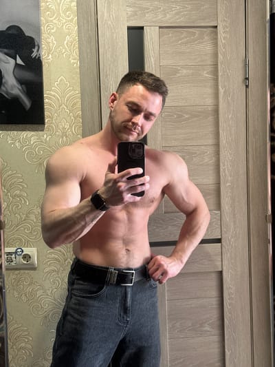 A shirtless man wearing jeans and a black watch is taking a selfie in a mirror. He is indoors with patterned wallpaper and a light wood-paneled door or wall behind him. A framed black and white image and an electrical outlet are also visible in the mirror's reflection. There is no verifiable indication of the city of Dobryanka, Russia.