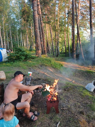 In Nepryakhinskoye Sel'skoye Poseleniye, Russia, a man wearing a black baseball cap and black shorts is sitting in a chair next to a campfire. He is shirtless and appears to be tending the fire with a green utensil. A young child with blonde hair, wearing a blue shirt, is standing in the foreground looking away from the camera. The scene is set in a forest clearing with tall trees and green undergrowth. There is a tent and a car visible in the background, suggesting a campsite. The ground is covered with dirt, grass, and pine needles. A white plastic bag and a brown paper bag are on the ground near the man. The lighting suggests it is late afternoon or early evening, with sunlight filtering through the trees.