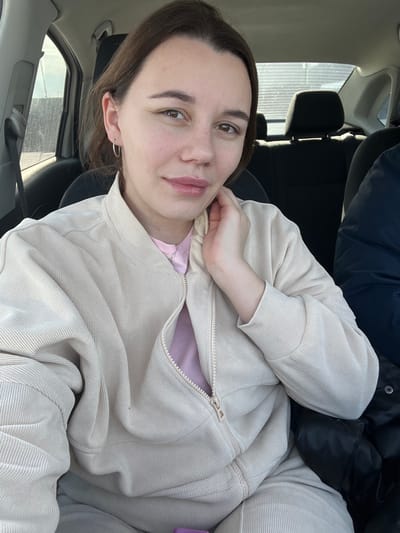 A single female subject is depicted inside a vehicle, at an unspecified location within the city of Shchepkin, Russia. She is positioned in what appears to be the front passenger seat, facing forward and looking directly at the camera with a slight smile. The woman has brown hair, visible eyes, and light skin, wearing small silver hoop earrings and a delicate silver necklace. She is dressed in a light beige, ribbed bomber-style jacket, partially unzipped, revealing a light pink shirt underneath. Her left hand is raised, with her fingers lightly touching her neck/chin area.
The vehicle's interior features black upholstered seats and headrests in the background. To the subject's right, a portion of another individual's arm or shoulder, clad in a dark blue or black puffy jacket, is visible. Through the vehicle's side window on the left, an outdoor scene is visible, featuring a light-colored vertical structure, possibly a fence or building wall, under natural daylight. A small purple object is partially visible at the bottom center of the frame.