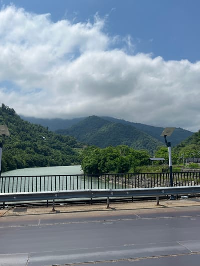 The image shows a scenic view of the Adjaristskali River in Georgia, with lush green mountains in the background. The river is calm and appears to be a light turquoise color. A bridge with a black metal railing spans across the river, with streetlights and a guardrail visible on the side. The mountains are covered in dense green foliage, and some clouds are present in the sky, suggesting it may be a partly cloudy day. The overall impression is one of natural beauty and tranquility.