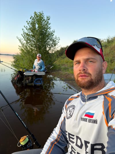 In Tarasovo, Russia, a man with a beard and a baseball cap is taking a selfie by the water. He is wearing a fishing shirt with the Russian flag and his name, "ГРОМОВ ЕВГЕНИЙ," printed on it. In the background, another person is fishing with multiple rods. The scene is set at dusk, with a tree and a body of water reflecting the sky. Fishing equipment, including rods and a tackle box, is visible.