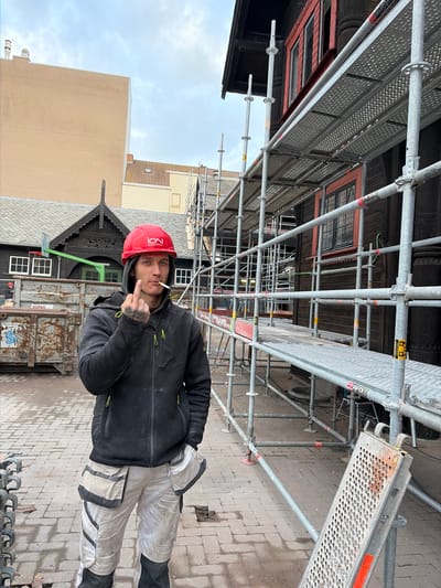 The image depicts a man wearing a red hard hat, a black jacket, and work pants, standing in front of scaffolding. He is holding a cigarette in his mouth and extending his middle finger. The location appears to be in Ostend, Belgium, as suggested by architectural styles and materials present. The scaffolding surrounds an older building with dark wood and window frames. A light-colored brick building is visible in the background. A large dumpster is positioned behind the man. The ground is paved with brick. The sky is overcast.
