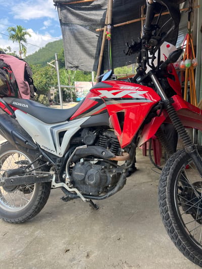 The image shows a red and white Honda XR190L motorcycle parked on a concrete surface. The motorcycle is positioned at an angle, with its front wheel on the right side of the frame and the rear wheel on the left. The motorcycle has "HONDA" branding on the seat and the engine casing. A black fabric covering is stretched over a bamboo structure above the motorcycle, suggesting it is parked under a makeshift shelter. In the background, there are trees, a hillside covered in vegetation, and a cloudy blue sky. To the right of the motorcycle, several colorful brushes or cleaning tools are hanging. The scene appears to be outdoors, possibly in a rural or semi-rural area, consistent with a location in Pac Ta, Vietnam. There are no people clearly visible in the immediate foreground or interacting with the motorcycle.