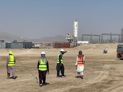 An elevated view captures an expansive, arid construction site under a clear, pale blue sky in Pir Dust, Afghanistan. In the foreground, four individuals, presumably male construction workers, are observed from behind or the side, wearing traditional attire augmented with high-visibility safety vests and hard hats. From left to right: one individual in a yellow hard hat and vest walks away from the viewer; a second wears a white hard hat, a grey head covering, a yellow vest, and holds a blue water bottle, walking forward with a dark measuring rope or line extending from a ground anchor near their feet; a third, in a white hard hat and yellow vest, faces right, actively pointing forward with his right arm; and a fourth, in a head covering and an orange vest, walks away from the viewer.
The midground consists of disturbed, light brown earth with scattered piles of sand or gravel. Numerous shipping containers, colored blue, brown, and white, are visible, likely serving as temporary site offices or storage. A prominent white concrete batching plant, marked with a "Z" logo on its silo, stands centrally. Further back, skeletal steel structures, resembling electrical transmission towers or a substation, are under construction. A person on a motorcycle traverses the dusty terrain from left to right in the mid-distance. The rear of a colorful, possibly decorated, truck is visible on the far right, with a yellow hose or cable extending from it onto the ground. The background features hazy, distant mountains blending into the horizon under the bright sky.