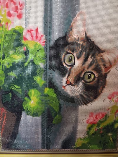 This is a close-up, diamond painting depiction of a calico cat peeking from behind lush green foliage, with pink flowers visible in the background. The cat's face is the central focus, with wide, alert green eyes and detailed tabby markings in shades of brown, black, and white. The painting style is characterized by small, colored beads or gems arranged in a grid pattern, creating a textured and mosaic-like effect. The environment appears to be an outdoor setting, possibly a garden or windowsill, with the cat interacting with the plants. The image is framed by a decorative border. The location of Комсомольский, Russia is not discernible from the image itself.