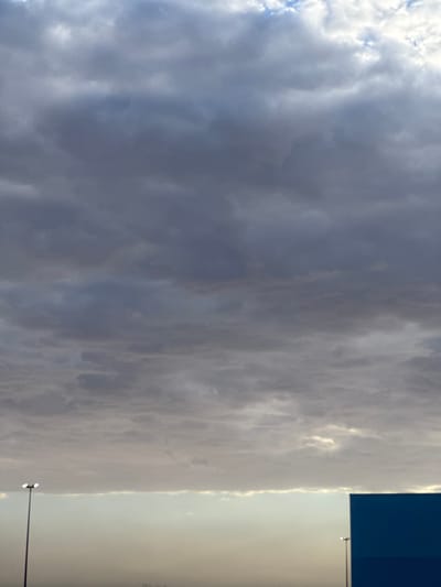 The image shows a cloudy sky over El Paso, Texas. In the lower portion of the image, a dark blue rectangular structure is visible on the right side. A streetlamp with two lights is positioned on the left side of the frame. The sky is filled with various shades of gray clouds, with hints of lighter sky peeking through in some areas. The overall atmosphere suggests an overcast or potentially stormy day.