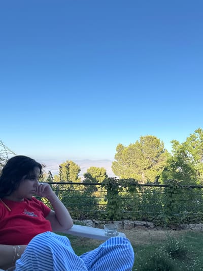 An outdoor scene in the Merom HaGalil Regional Council, Israel, shows a seated individual with dark hair, oriented towards the left frame edge. The person wears a red short-sleeved shirt with a white graphic and blue and white striped pants. Their left hand is raised, fingers lightly touching the chin area, displaying a ring on the left ring finger and bracelets on the wrist. A yellow object is visible near the left shoulder. The individual is seated in a white outdoor chair, on the right armrest of which rests a clear glass containing liquid. Behind the chair, a dark-colored metal fence is partially covered with green climbing vines. Beyond the fence, a natural landscape unfolds, featuring numerous green trees, including tall conifers, with distant, hazy blue-grey hills under a clear, bright blue sky. The immediate foreground on the ground consists of green grass interspersed with small white flowers. The scene is illuminated by bright daylight.
