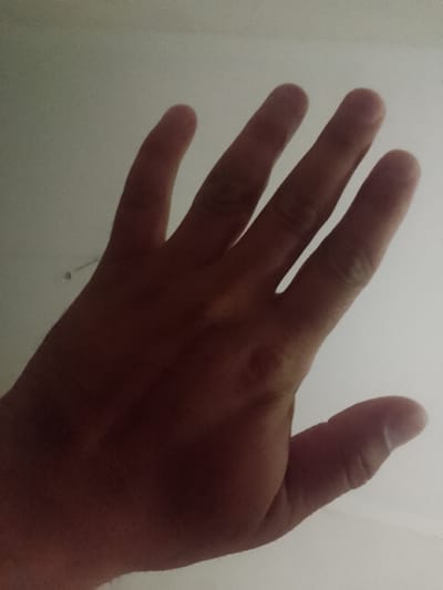 A close-up, low-angle shot shows a hand with fingers spread open, palm facing upwards against a white, slightly textured background. The hand is casting a shadow, suggesting a light source from above. The lighting creates a subtle gradient of light and shadow across the skin. The image is taken indoors, possibly in a home or office, in Махамбет ауылдық округі, Kazakhstan, though the specific urban context is not evident from the visual alone.