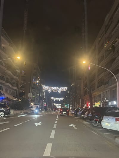 An angled view of a brightly lit street at night, with decorations hanging over the road. Cars are parked along the side of the road and driving on the street. Streetlights are evenly spaced and illuminate the scene, while the arrow sign on the road indicates the direction of travel. Buildings line either side of the street, adding to the urban landscape.